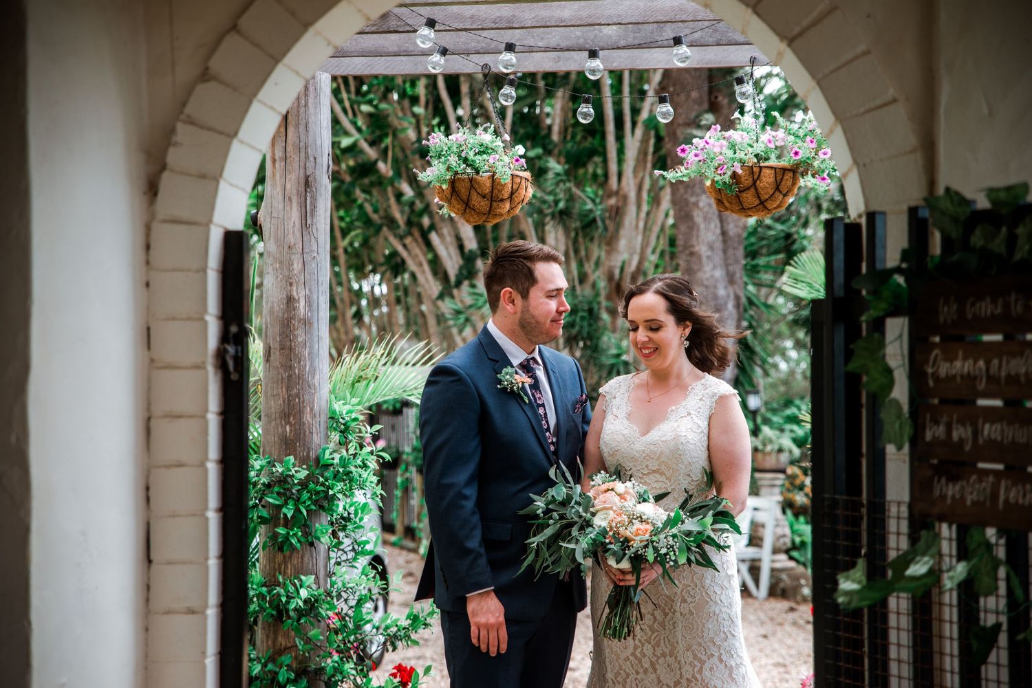 Wedding couple standing under white stone archway surrounded by lush greenery.