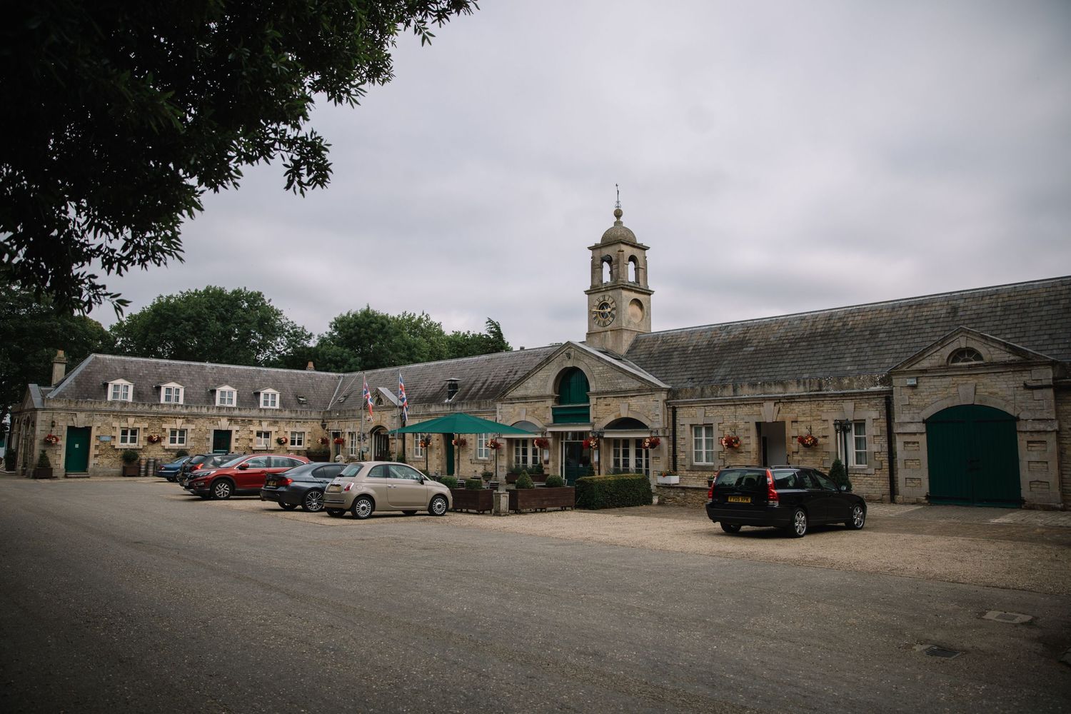 A historic town square with stone buildings, clock tower and parked cars on a cloudy day.
