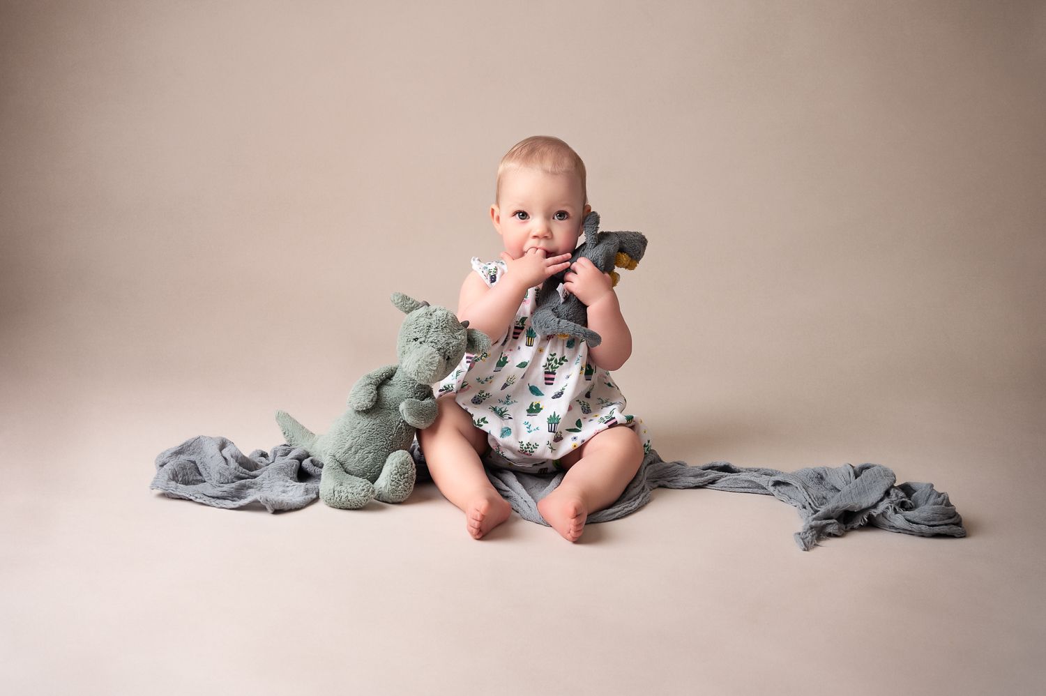 Baby sitting with stuffed dinosaur toys on beige backdrop during portrait session.