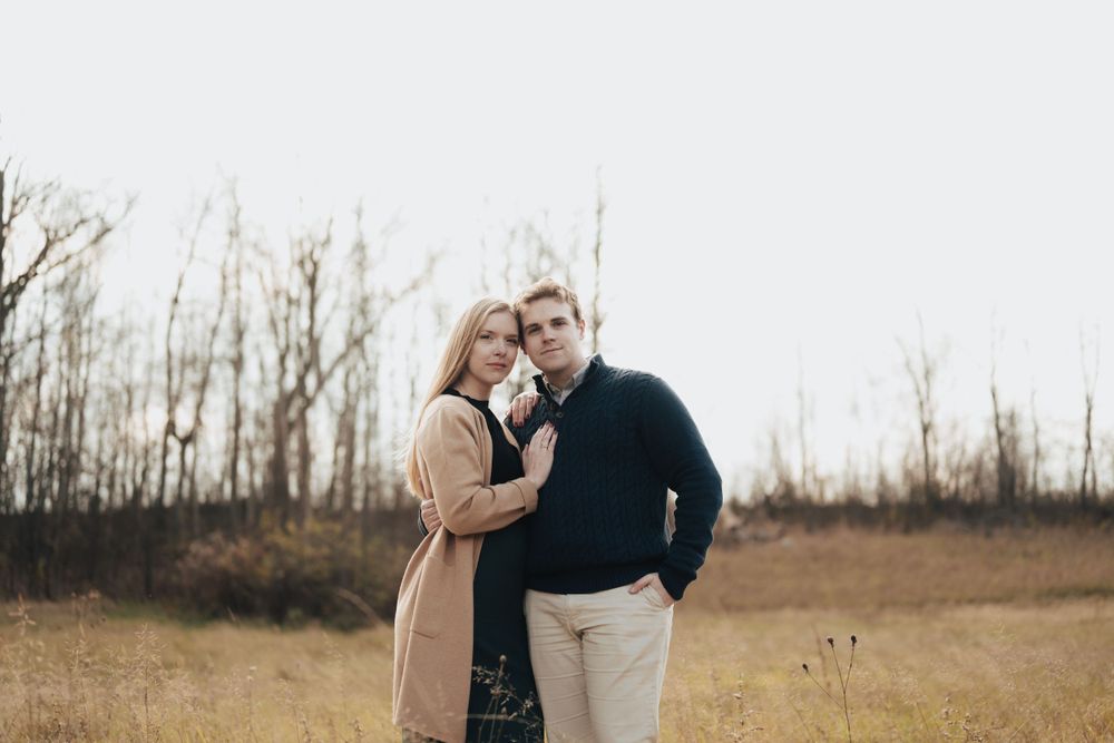 Couple posing together in an autumn field with bare trees in background.