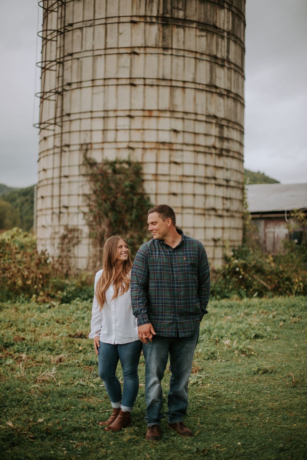 Couple holds hands smiling at each other beside a rustic farm silo on an overcast day.