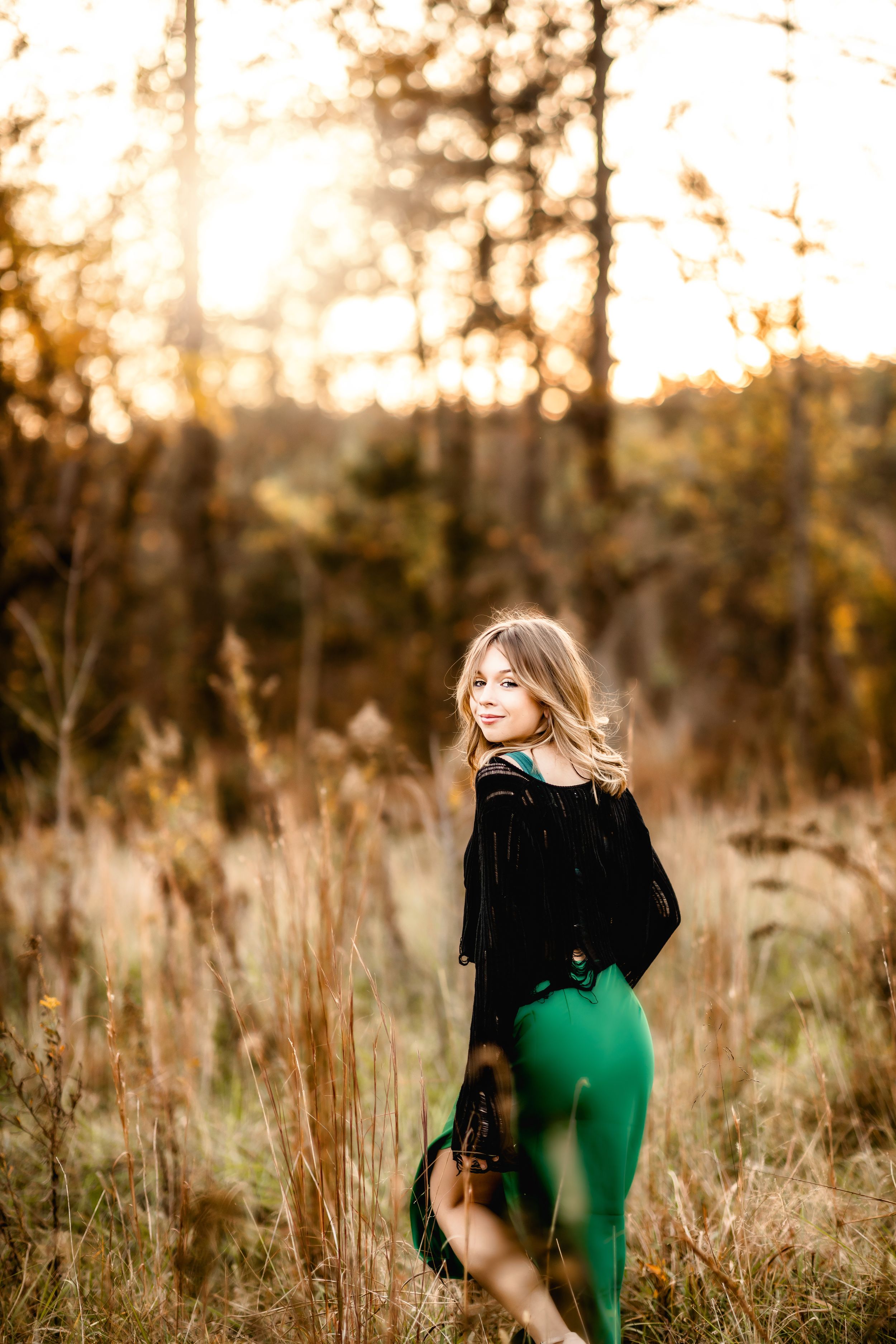 Woman in black velvet top and green pants poses in golden autumn field.