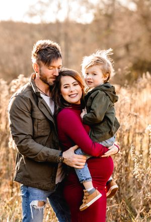 Family of three embracing outdoors with mother in red dress and autumn scenery.