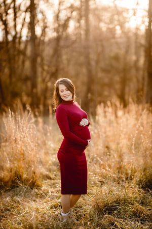 Maternity portrait of woman in red dress standing in golden field at sunset.