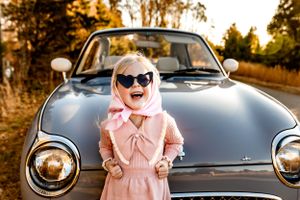 A playful pose in front of a classic car wearing a pink dress and sunglasses.