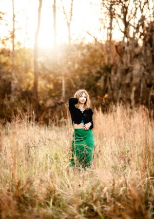 Artistic portrait of a figure in a green skirt standing in tall golden grass at sunset.