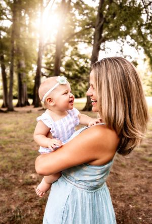 A joyful outdoor portrait of a baby and parent smiling together in a sundress against sunlit trees.