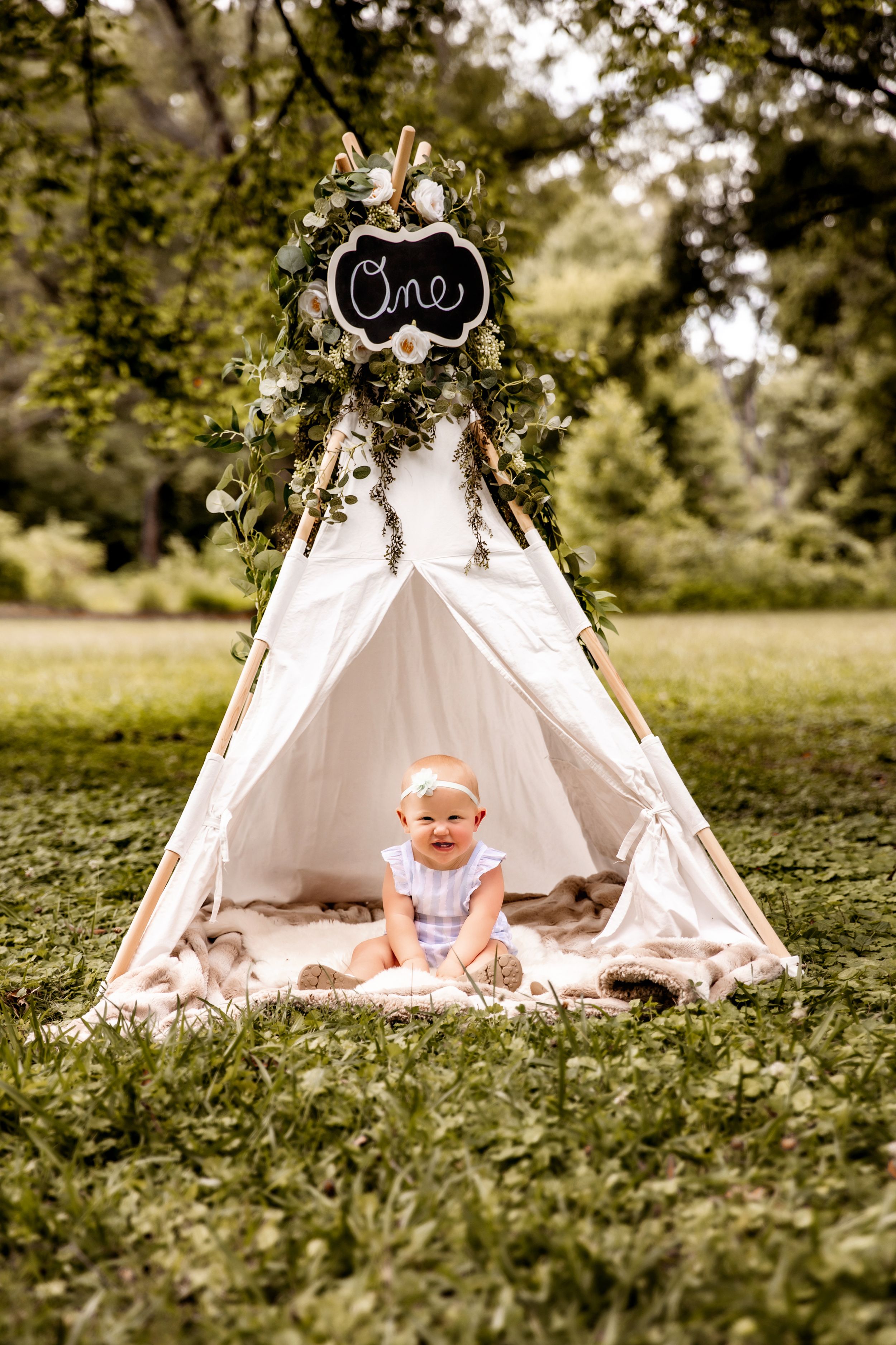 Baby sits inside decorated white teepee tent with 'One' sign during outdoor photo session.