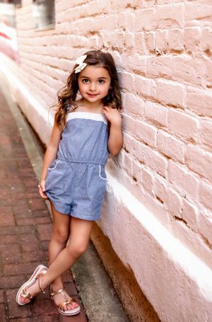 Young child in blue romper posing against pink brick wall with flower in hair.