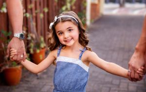 Child in denim dress smiling while holding hands in urban walkway.