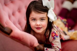 Close-up portrait of a child wearing a white bow on pink tufted furniture.