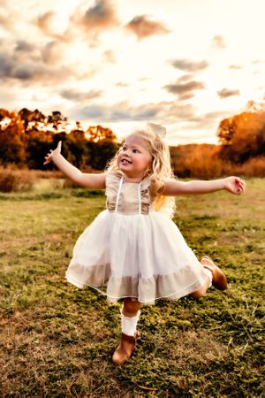 Young child in white dress twirling in field during golden sunset.