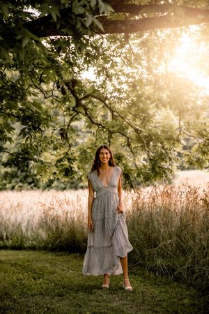 A gray maxi dress stands elegantly in a sun-drenched field with trees casting dappled shadows.