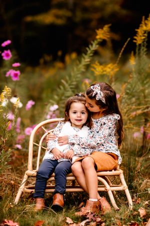 Two children sitting on a rattan bench among wildflowers sharing a sweet moment together.