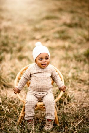 A happy baby in a knitted outfit and white beanie sits in a wooden chair outdoors.