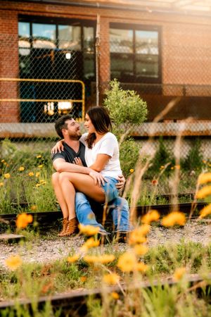 Romantic couple sitting among wildflowers near brick building during golden hour.