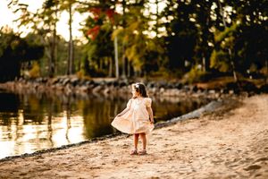 Small child in pink dress exploring sandy beach at sunset with lake and trees in background.