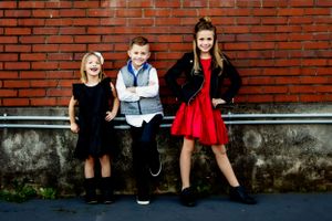 Three children posing against red brick wall wearing dressy outfits.