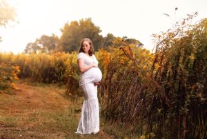 A maternity portrait of a woman in white dress standing on a rustic path at golden hour.