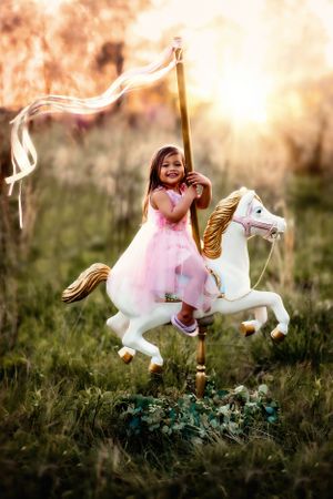 Child in pink dress rides decorative carousel horse in dreamy sunset setting.