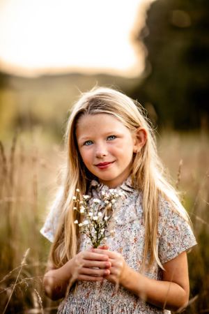 Child in floral dress holding wildflowers in golden sunset field.