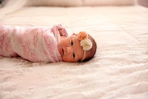 Newborn baby wrapped in pink swaddle lying on cream bedding with floral headband.