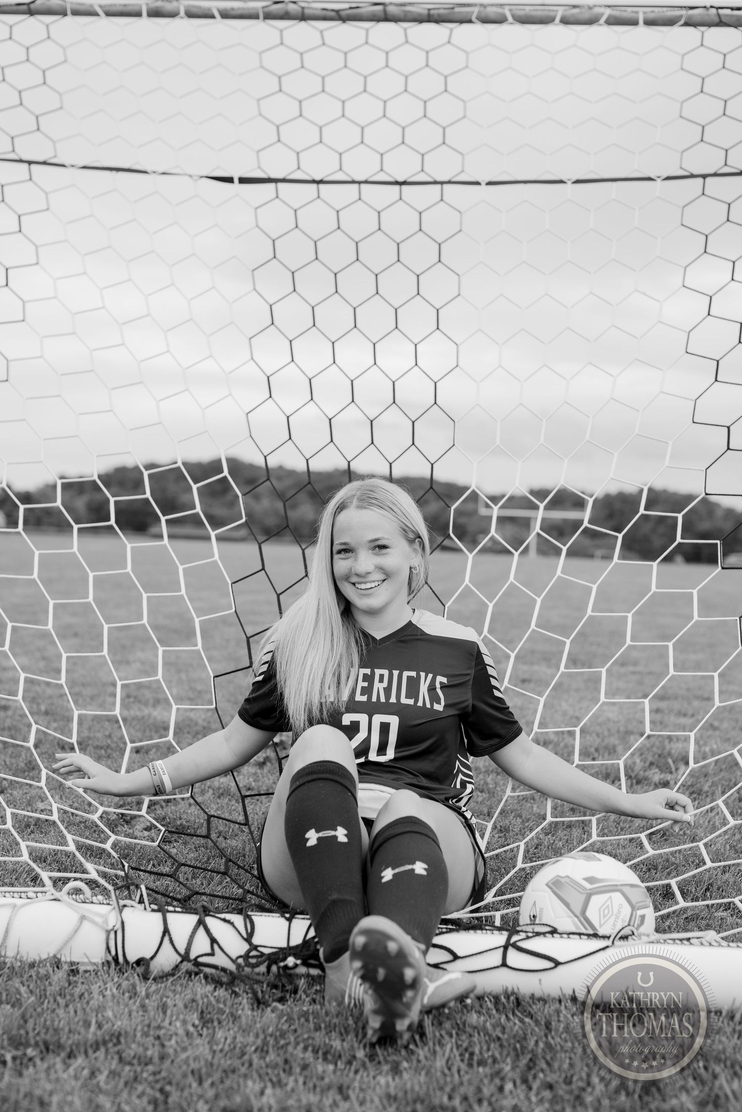 Black and white photo of soccer player sitting in goal net with ball and athletic gear.