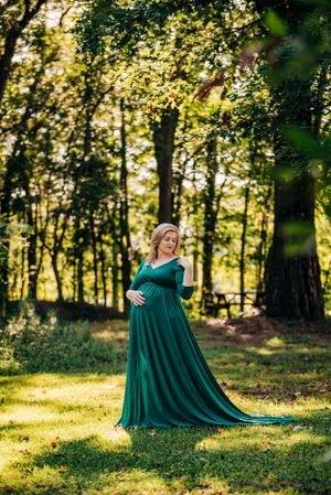 Maternity portrait of woman in emerald green gown standing in sunlit forest.