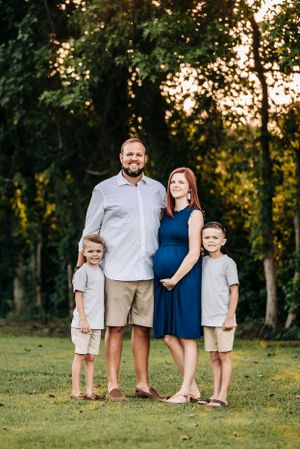 Family of four poses together outdoors with expecting mother in blue dress.