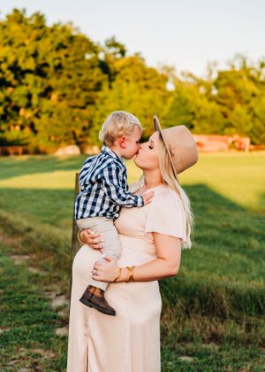 Mother in beige dress and hat sharing tender moment with toddler outdoors at sunset.