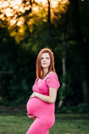 Expectant mother in pink outfit poses against wooded backdrop at sunset.