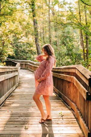 A maternity photo of someone in a pink dress twirling on a wooden boardwalk surrounded by forest.