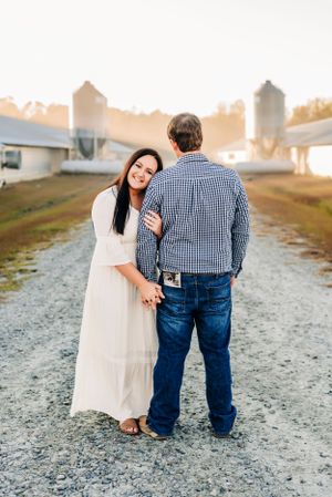 A romantic countryside photoshoot featuring a couple posing on a gravel path with grain silos in the background at sunset.