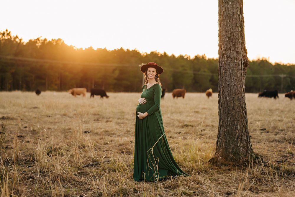 A woman in a flowing green dress stands between trees at sunset with cattle grazing behind her.