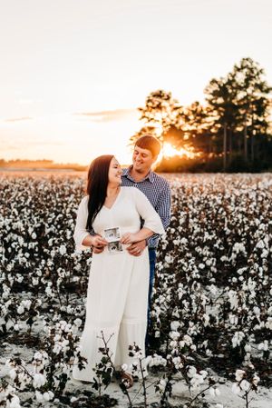 A couple embracing in a blooming cotton field during golden hour with the sun setting behind pine trees.