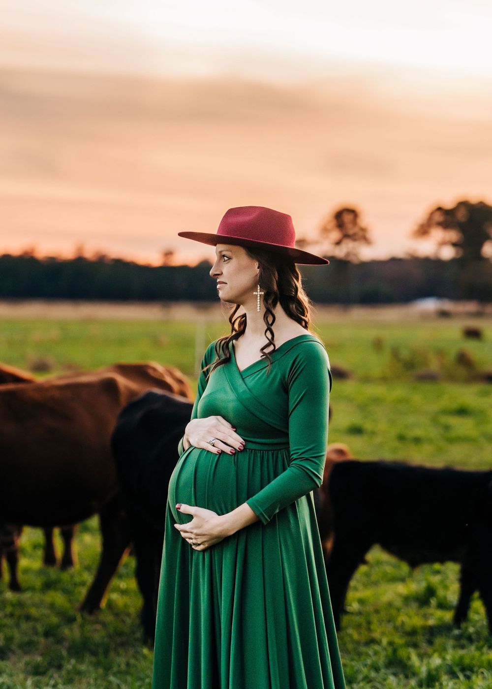 A woman in emerald dress and burgundy hat stands in field with cattle during sunset.