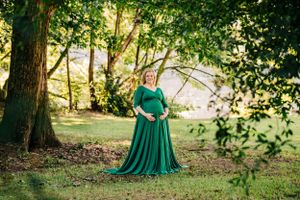 Woman in flowing green dress poses for maternity photo in wooded setting.