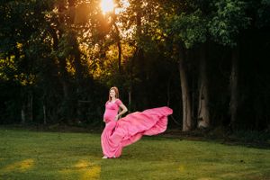 Woman in flowing pink dress poses dramatically at sunset in wooded setting.