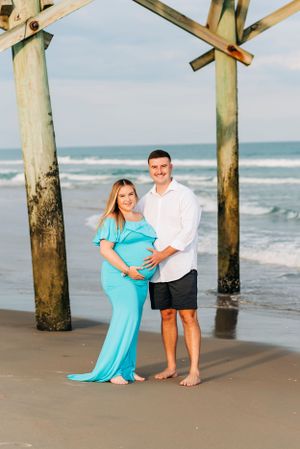 A beachside maternity photo under wooden pier posts with waves in the background.