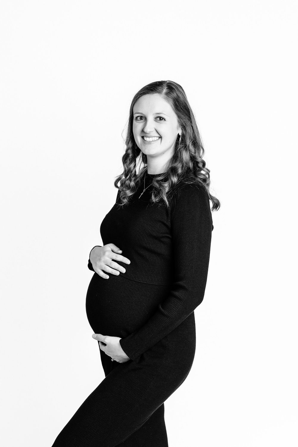 A woman in black dress poses for maternity photo against white background.