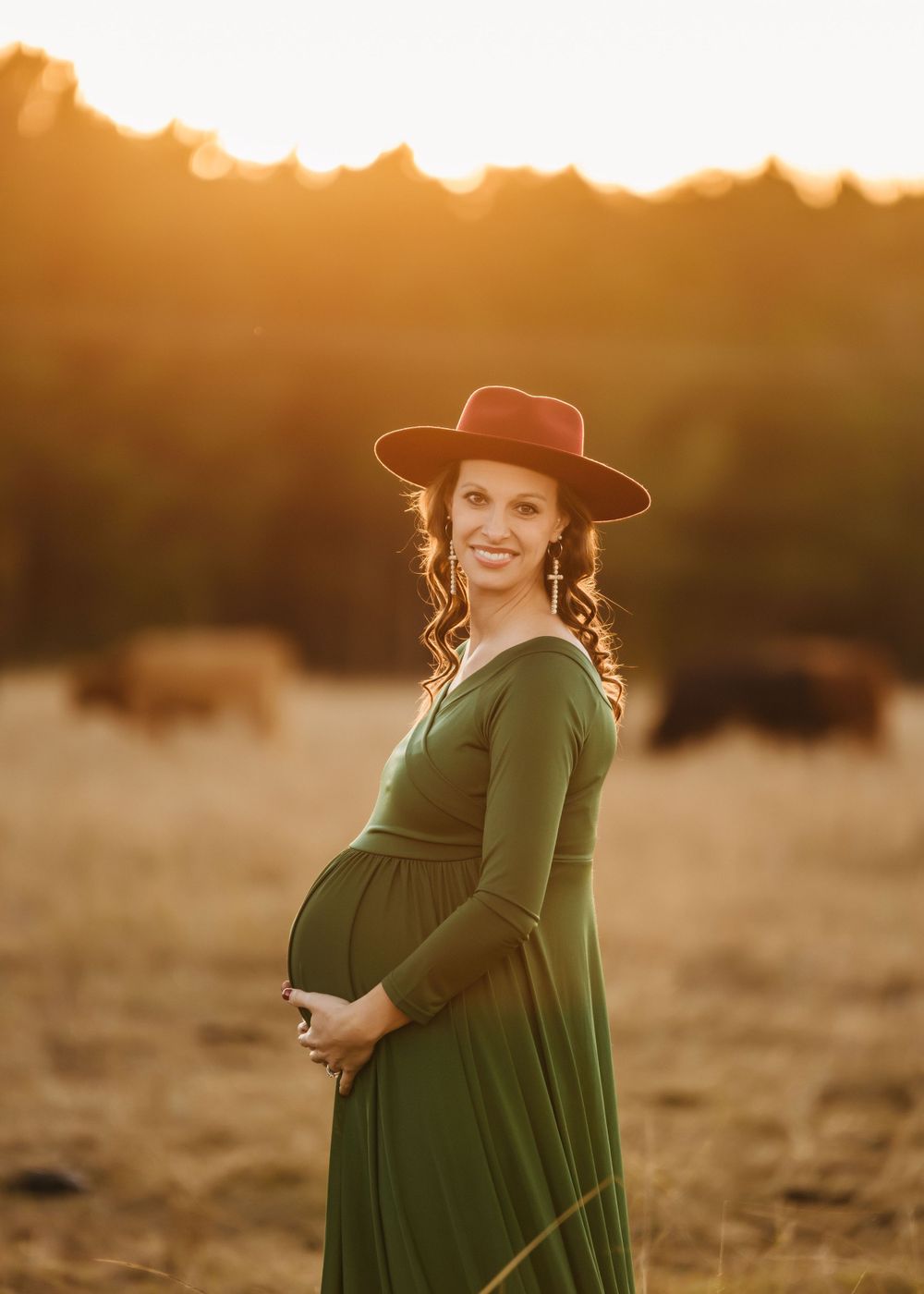 A woman in green dress and brown hat poses in a field during golden hour.