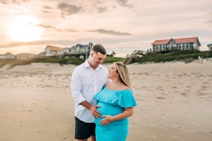 A couple poses for maternity photos on the beach at sunset with coastal homes in the background.