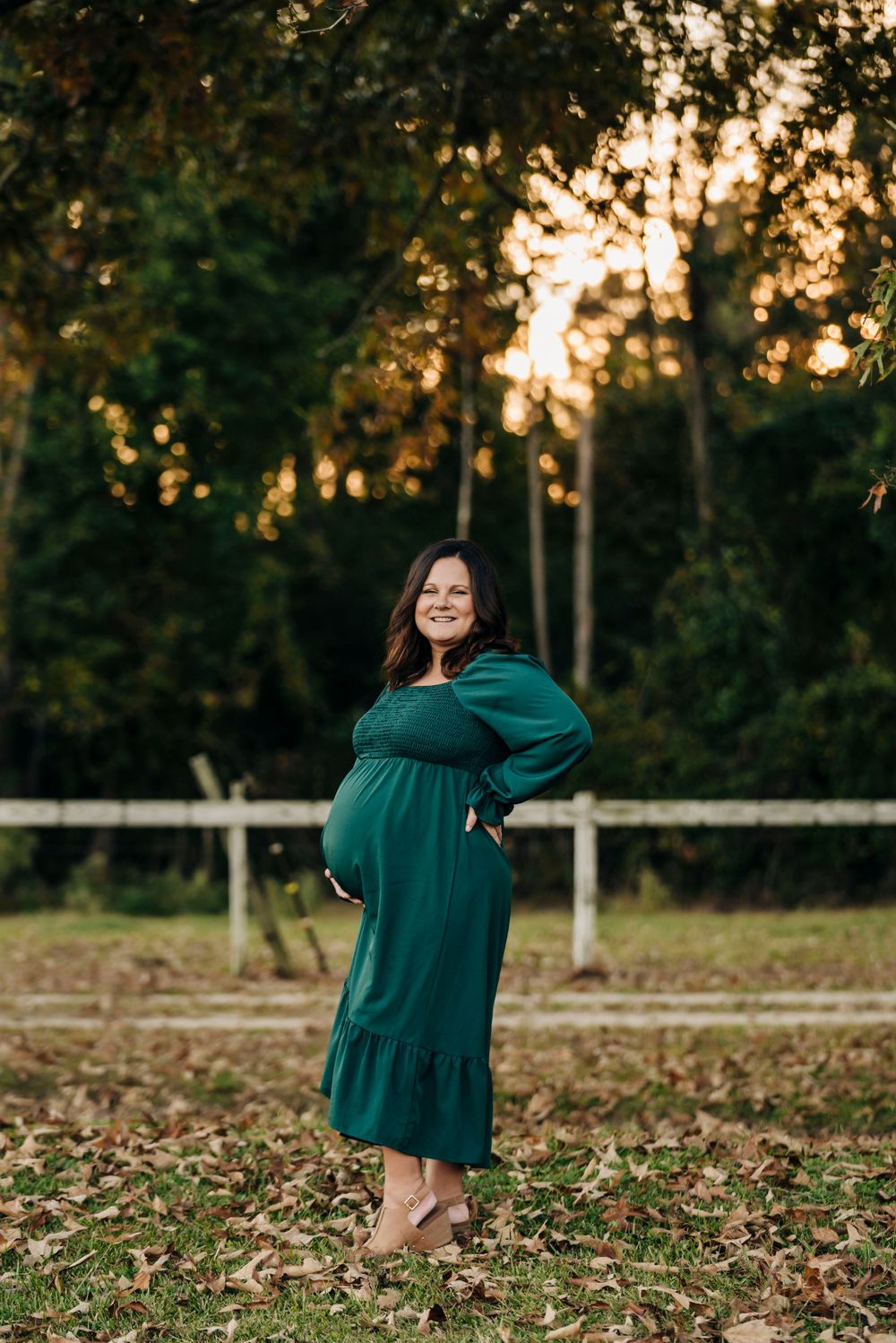 A woman in emerald dress poses in front of white fence during golden hour.