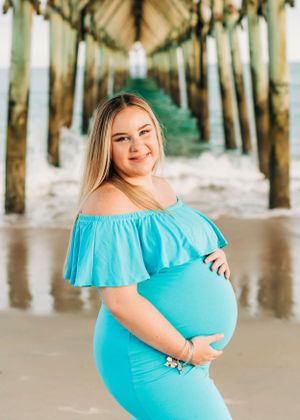 A maternity portrait in a turquoise dress under a beach pier with waves visible in the background.
