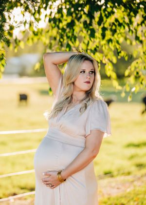 Maternity photo of expecting mother in white dress posing under sunlit tree branches at golden hour.