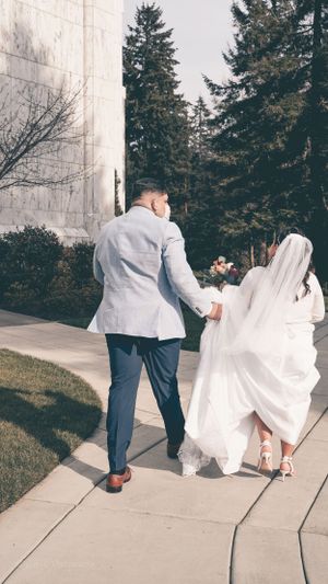 A couple walking along a stone pathway beside an architectural wall and evergreen trees.
