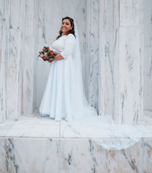 Bride in elegant white gown holding bouquet against marble wall backdrop.