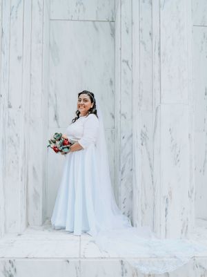Bride in elegant white dress and long train poses against a light textured wall while holding a floral bouquet.
