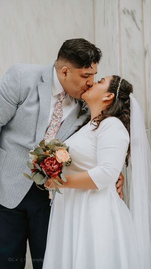 Newlyweds share a romantic kiss against a white wall, with bride in flowing gown and groom in light gray suit.