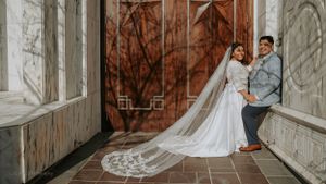 Wedding couple poses playfully by wooden door with bride's long lace veil trailing behind.